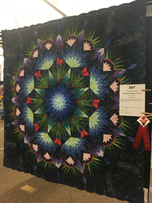A quilt done in the style of a broken carpenter’s star with greens, blues, and pink highlights. Lovely natural and butterfly highlights