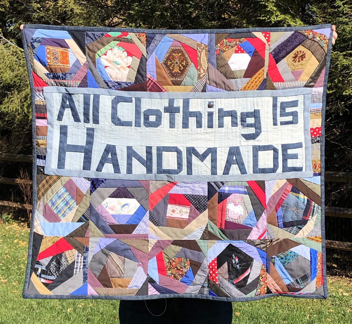 A quilt made of reused textiles, pieced as squares in rows of improvised blocks. Each non-lettered row has 5 blocks. There is an upper row, then an upper middle section in dark and light denim that reads: All Clothing is Handmade. Then two lower rows. Colors tend toward reds, browns, blues, and a lot of purple.