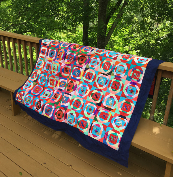 a full view of the quilt draped over my porch railing. it is a square with 100 blocks each with its own unique and colorful little critter. prominent colors are red, pink, light blue, dark blue