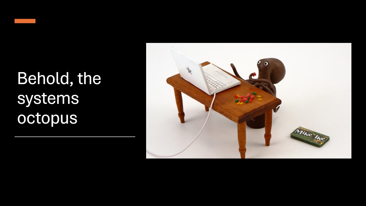 A slide showing a handmade miniature octopus sitting at a computer and eating tiny jelly beans. The slide says: Behold, the systems octopus