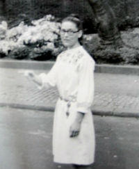 A photograph of Mom in college. Black and White. She has thick black glasses on, a white dress, and is pointing off-camera.