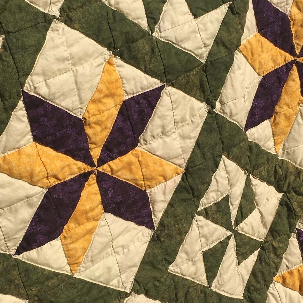 A closeup which shows that the background color fabric is a tan shade made even more golden by the setting sun. The hand-stitching is “in the ditch” following the outside of the stars, the pinwheels, and the border. The stars also have sitching on one side of each inner seam.
