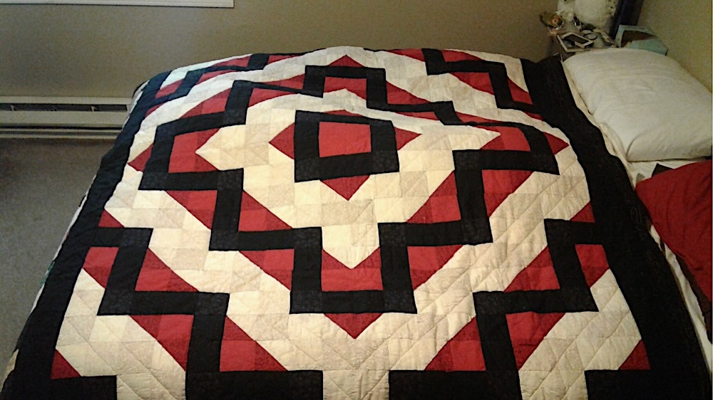 A quilt lying on a bed. It has a background of red and cream diamonds which alternate out from the center, resembling a log cabin. Each red section has line of black blocks which follow the diamond but zig-zag jaggedly across it. While the sections might initially appear to be one color each, there are subtle variations in each color palate. 3 shades of cream, 2 of black, and 2 of red.