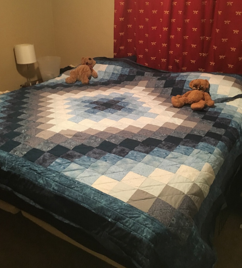 The finished quilt lying on a bed, photographed at an angle. Two teddy bears are perched on top of the pillow.