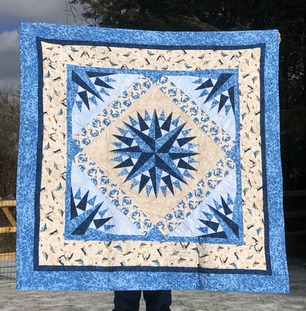 A quilt done in the Mariner’s compass pattern. The center compass has medium and dark blues in a couple of layers. The medium blue is like waves. There’s then cream background to make a square, a border of crab fabric. the same compass pattern is in additional corners with light blue backgrounds and then a border of shore birds… followed by a thin border of dark blue and a fuller one of medium blue fabric. It’s layers of blues and creams. The picture is taken outside with beautiful sunshine on it.
