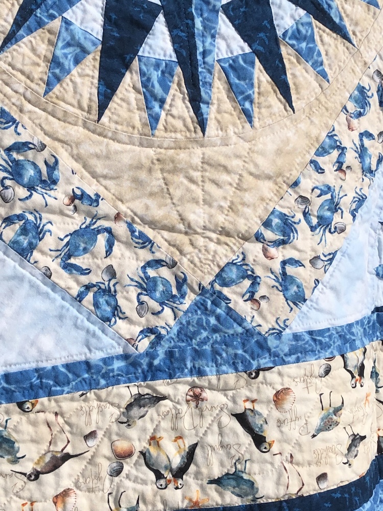 A detailed picture of a quarter of the quilt front which shows quilting details. Compass spikes are outlined. The fill around the compass radiates from the corner toward the circle. There’s a twisting pattern through the crab and bird borders.