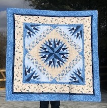 A quilt done in the Mariner's compass pattern. The center compass has medium and dark blues in a couple of layers. The overall pattern is mostly medium and light blues and creams with dark blue accents.
