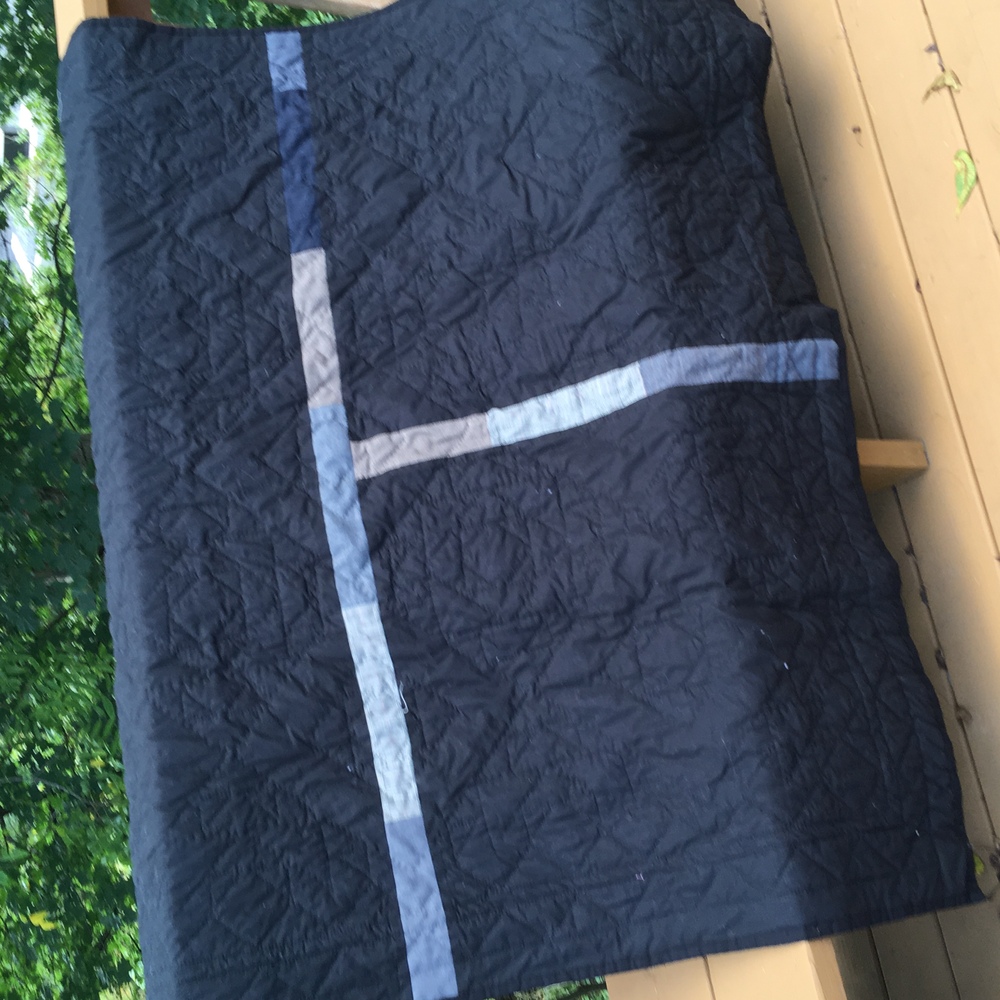 The quilt hung on the deck to show its back. The back is black but a skinny T cuts it into two medium and one large segment. The T is pieced from long, thin strips cut off the hems of men’s trousers. You can see intense diamond quilting.