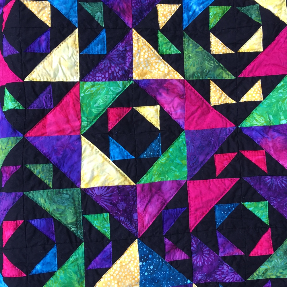 An outdoor closeup of the quilted top focused on just one diamond’s pattern with a black diamond set in batik square set in black diamond set in batik diamond. Black quilting stitches are visible in batik squares.