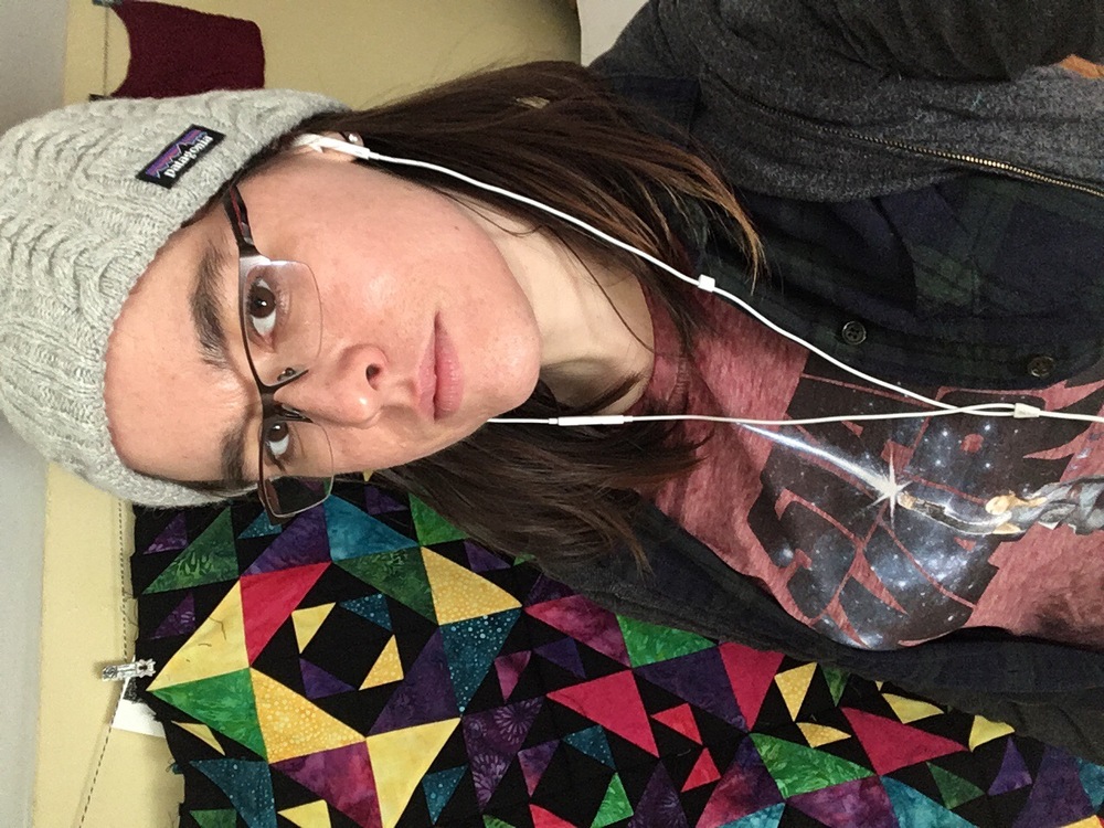 A selfie of me, with shoulder-length brown hair and skinny-framed glasses. I’m in front of the quilt, wearing a hat and looking at the camera with a slight smile.