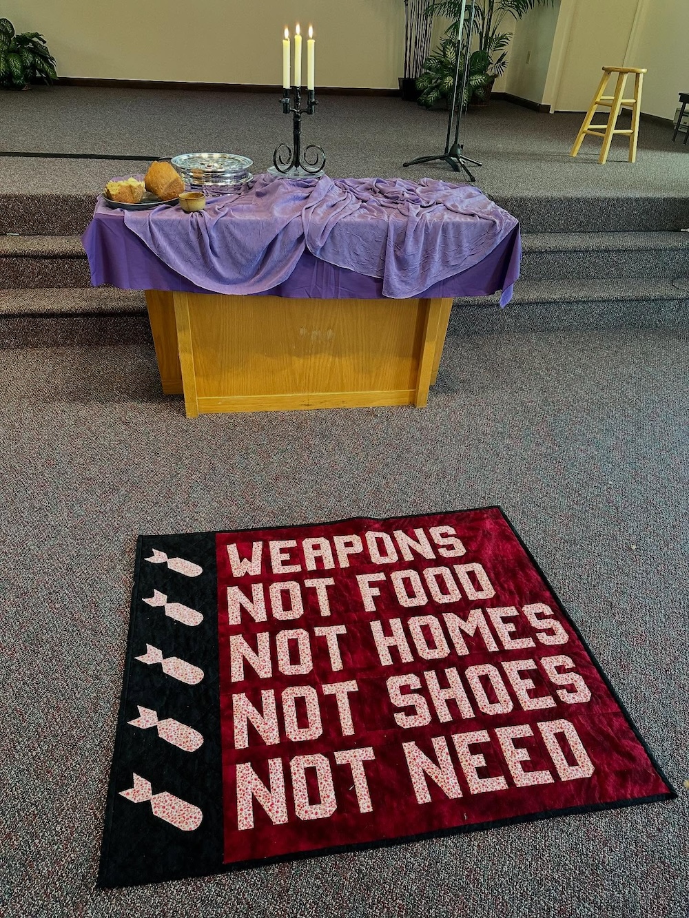 The quilt lies on the floor in front of a communion table. A trio of lit candles are in a stand on the table made of three bayonets from WWI, a gift from a German church.