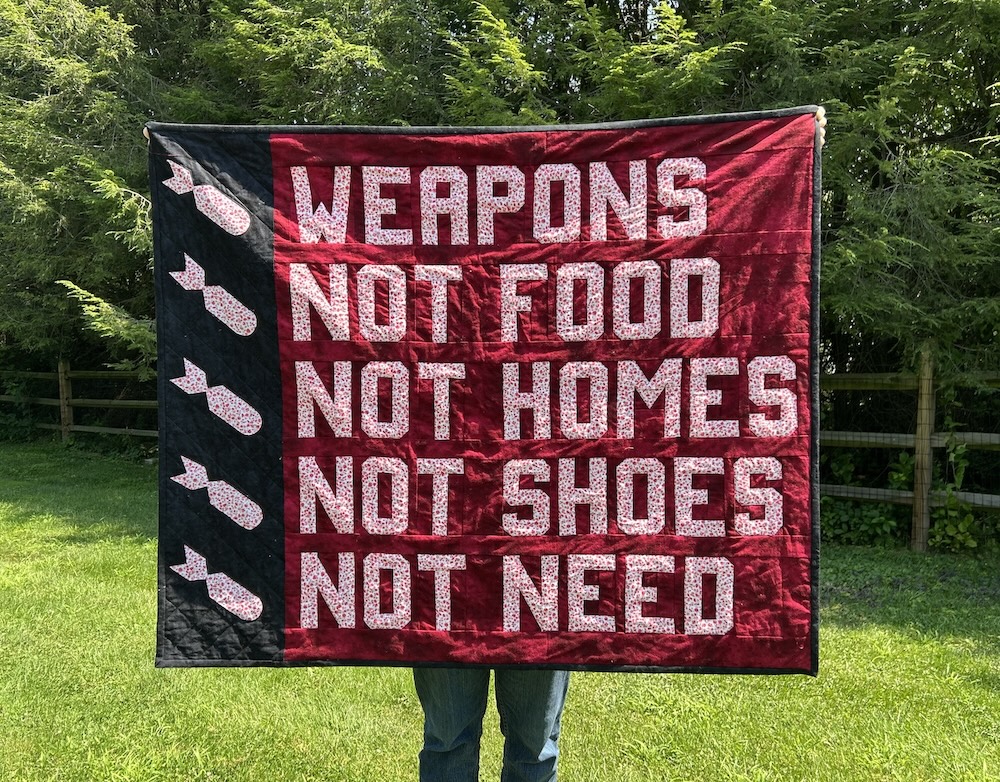 A throw-sized quilt being held in the sunlight. The words Weapons Not Food Not Homes Not Shoes Not Need are pieced in a white cotton fabric with little red flowers on a deepl and blood-like red background. On the left side, punctuating each line like bullet points, are appliqued bombs, again in white fabric with red flower prints on a black background.