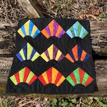 A mini quilt with colorful fans on a back background. The fans are bright, bold batiks in oranges, greens, yellows, blues, and reds, with a black arc piece at the fan base.