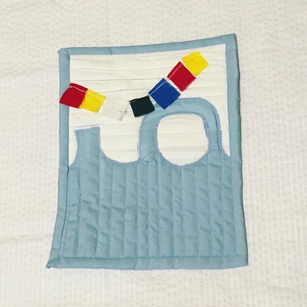 A small quilt which appears to show the side of a house (with white siding) and a rather basic light blue fence which comes up in a point on the left side, then has a circle effectively connecting two points on the right. A row of colorful squares is string together and hanging across it. The execution is a bit rough, experimental vs. refined