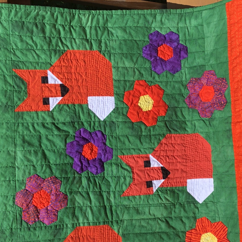 A partial closeup of two foxes and a subset of flowers. The foxes have tighter quilting than the background and the flowers are only outlined.