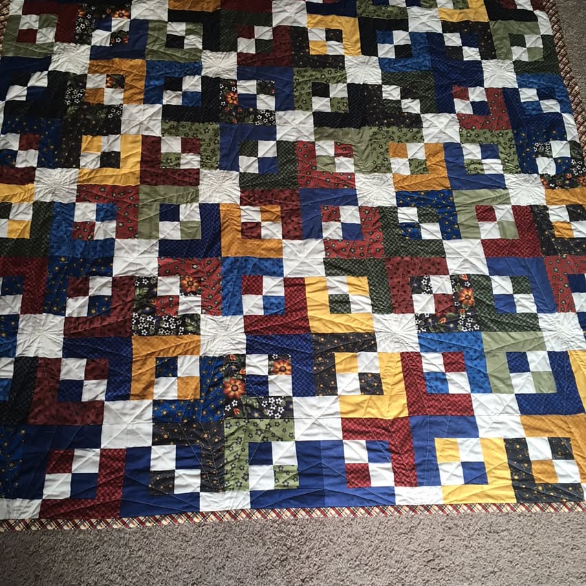 The full quilt laid out on the carpet in a darker room. The small and medium-sized white blocks running in a diagonal pattern contrasts with medium and dark fabrics of the blocks between them.