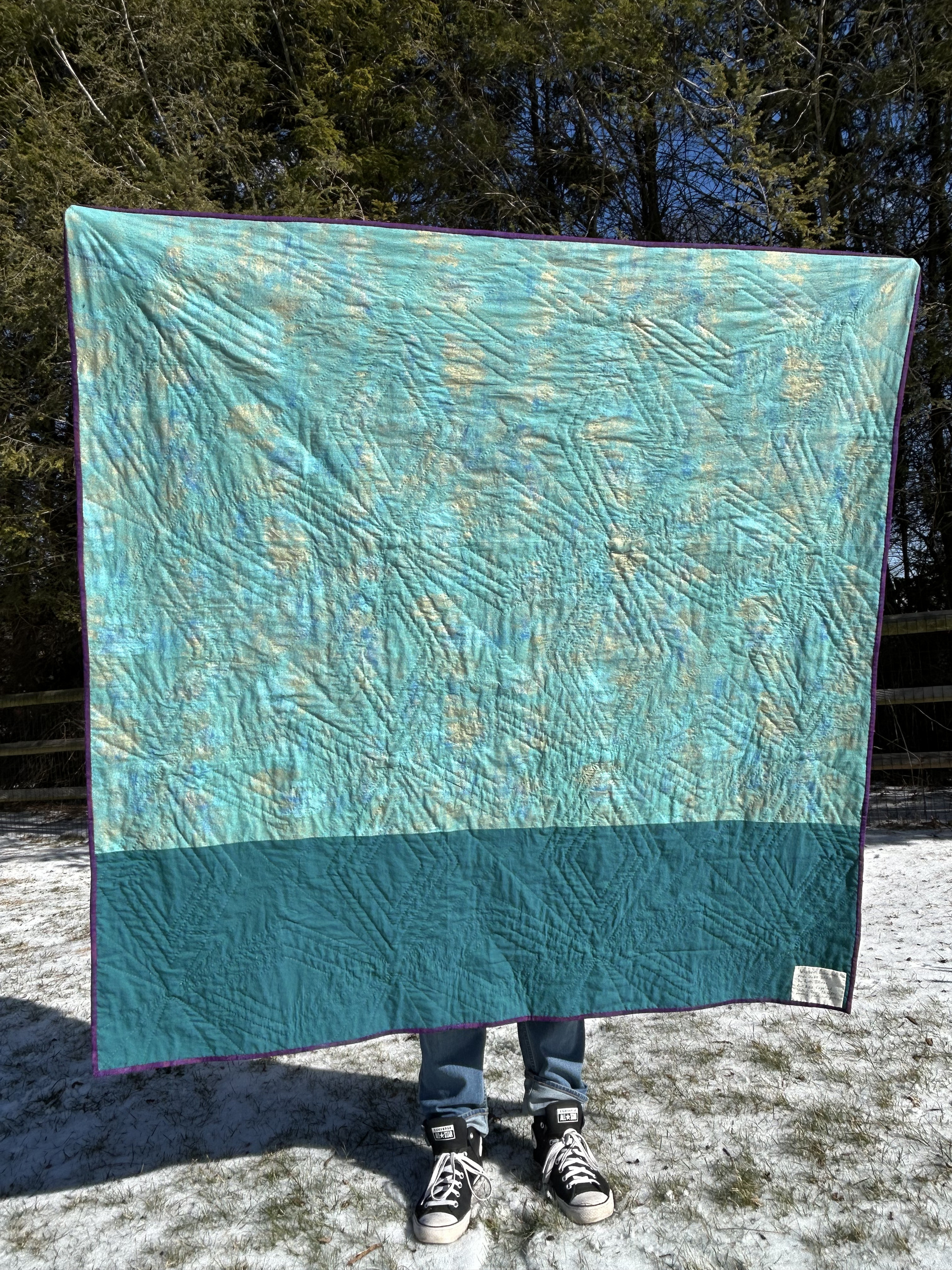 Back of the quilt. The fabric is mostly teal with gold dusting for emphasis. The quilting reflects diamond shapes of the front, connected by radiant diagonal lines. The bottom band of the quilt back is about one foot of dark teal