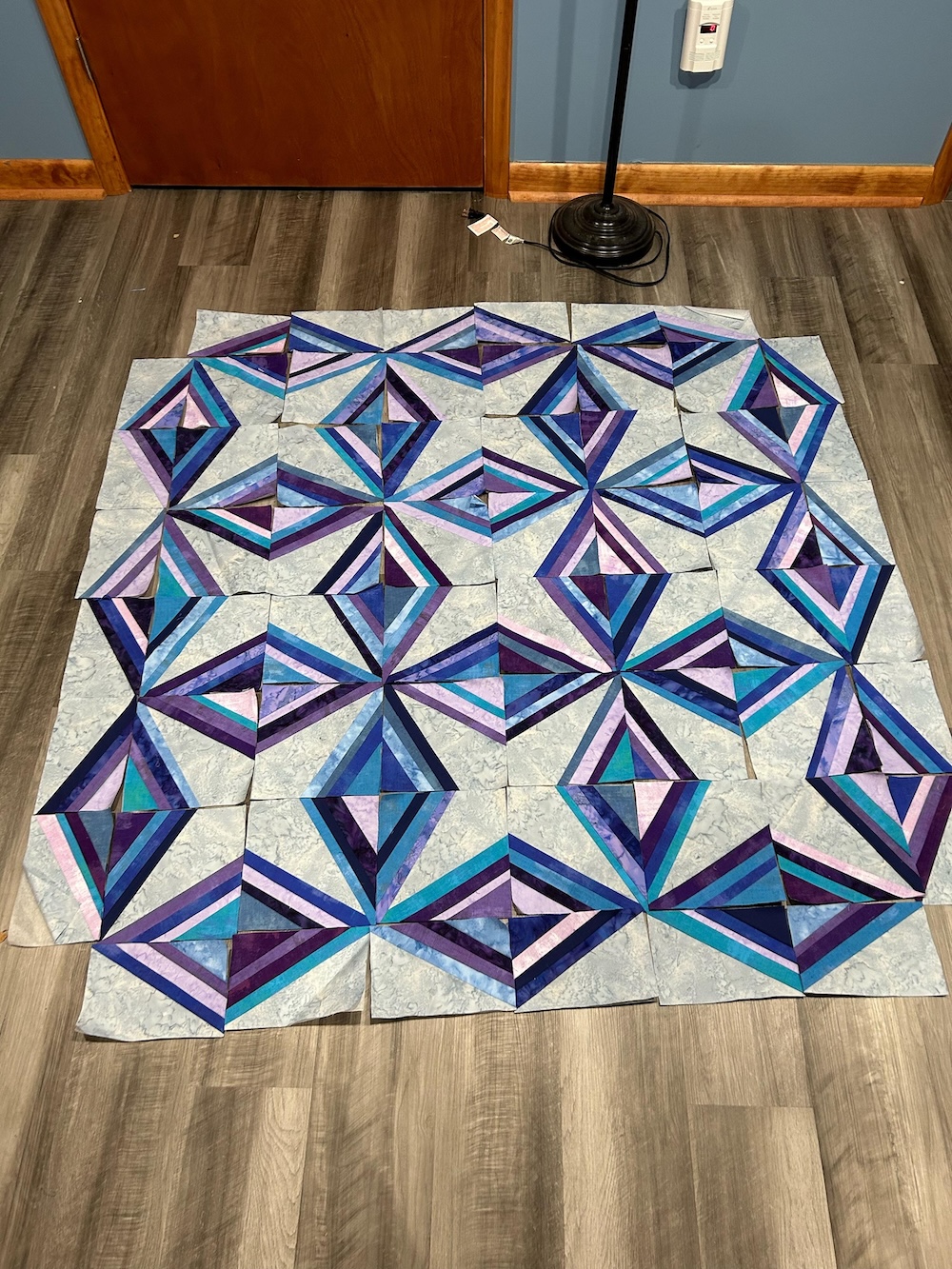 Quilt blocks laid out on the floor. Each block has a wedge of gray in the center and two wedges of created through stripes. These come together to create a faceted impression