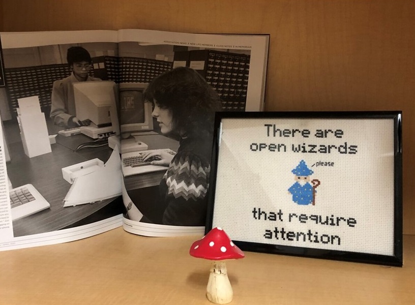 A photograph of a shelf with the image of women using LIAS terminals in a magazine spread and a cross-stitched error message: There Are Open Wizards That Require Attention