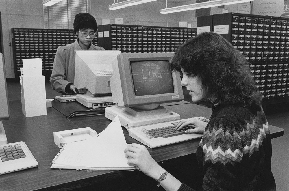 A slide showing two young women, one Black and one white, using terminals in the card catalog room. The LIAS welcome screen is on the terminal we can see.