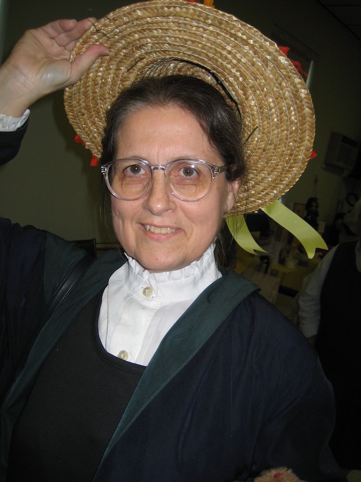 A woman in her late 50s with big frame glasses is smiling and removing a straw hat that has a ribbon on it. She looks very much like me.
