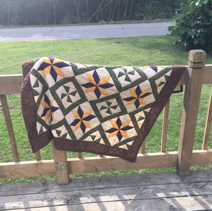 A throw-size quilt hangs on a porch railing in the evening sun. It's made up of 9 large motif blocks set on point as diamonds with small motif blocks between them and out to the brown broder. The large blocks are stars with alternating purple and orange points, like a striped flower. The alternating blocks are green pinwheels with a green border. The effect is a bit like a neatly-planted garden with green separating the flowers.