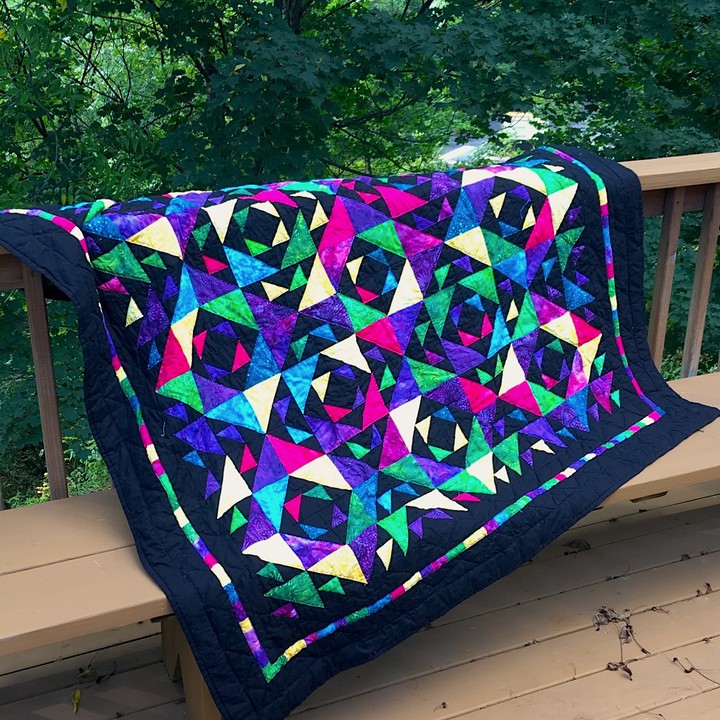 A large square quilt draped over a porch railing. The quilt is bright jewel-toned batiks in pinks and blues and greens and yellows especially on a black background. Bigger triangles form diamond shapes around central black diamonds that are set in multi-colored batik squares.