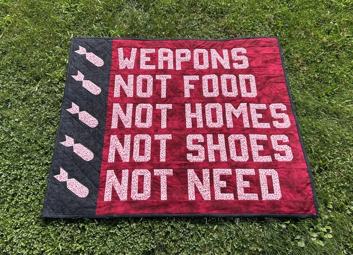 A throw-sized quilt lying in the grass. The words Weapons Not Food Not Homes Not Shoes Not Need are pieced in a white cotton fabric with little red flowers against a contrasting deep and blood-like red background. On the left side, punctuating each line like bullet points, are appliqued bombs, again in white fabric with red flower prints on a black background. Black quilting on the black background shows a cross-hatched pattern, evoking a chain-link fence.