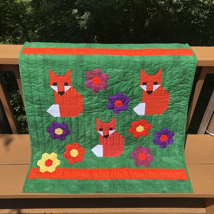 A green quilt with three orange foxes on it. There orange, purple, and yellow flowers appliqued onto the quilt. The quilting is mostly done in straight, broad lines with some finer quilting on the foxes themselves. One of the flowers overlaps an orange border a little for whimsy.