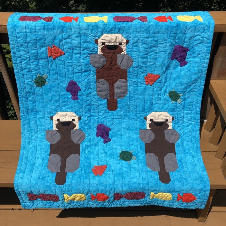 Three pieced otters float on a variegated blue background evoking water. Borders above and below them are pieced fish in purple, orange, and yellow. A set of appliqued 3D fish and turtles swim around them.